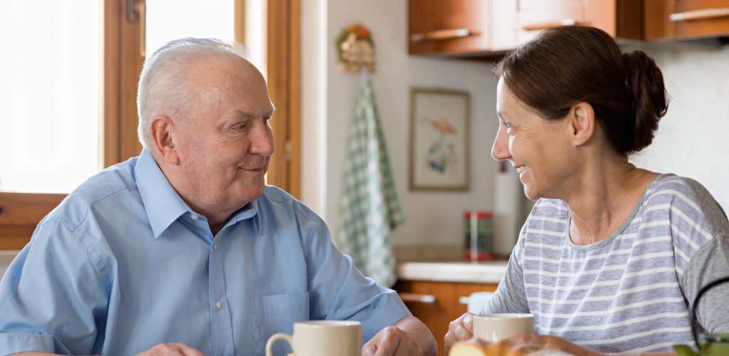 Foto de una mujer de mediana edad junto a un hombre mayor. Ambos están sentados en la mesa de una cocina y toman té mientras se miran y sonríen.