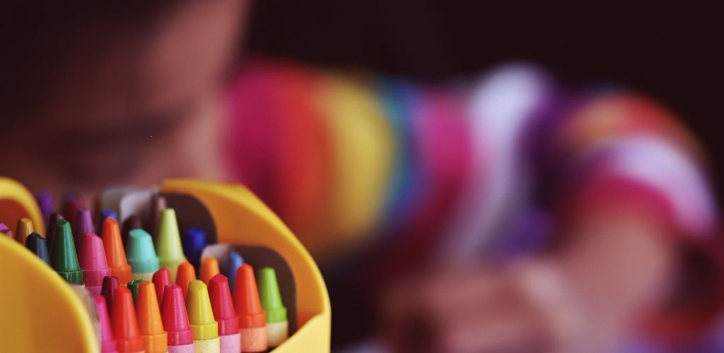 Foto de lápices de cera de colores. Detrás, borrosa, se ve una niña pintando con lápices.