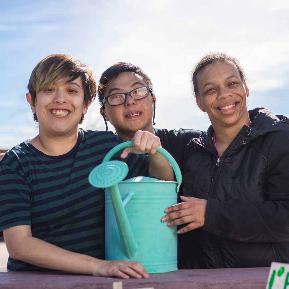 Foto de tres personas con discapacidad intelectual sonriendo. El joven del medio sostiene una regadera.