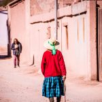 Foto de una mujer mayor con sombrero y bastón, caminando de espaldas por una calle del norte de Chile que es de tierra. 