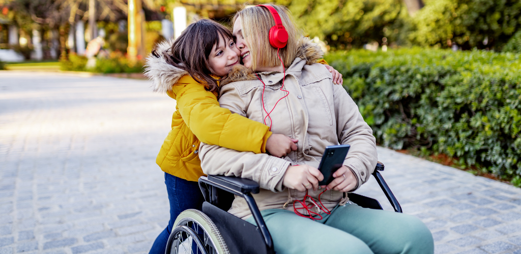 Foto de una mamá en silla de ruedas junto a su hija pequeña que la abraza.