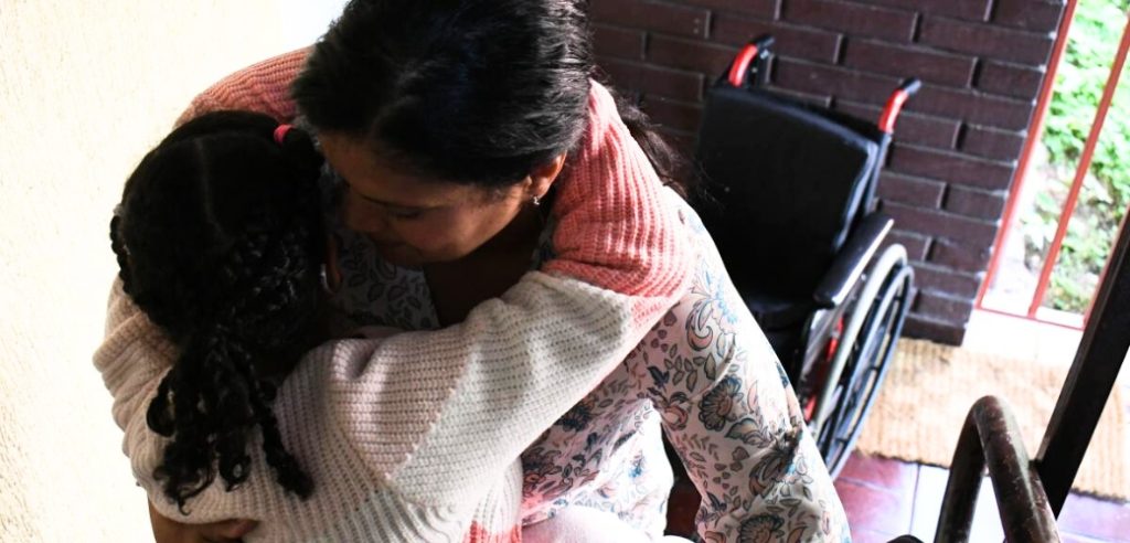 Mujer cuidadora sosteniendo en sus brazos a una niña. En el fondo se ve una silla de ruedas.